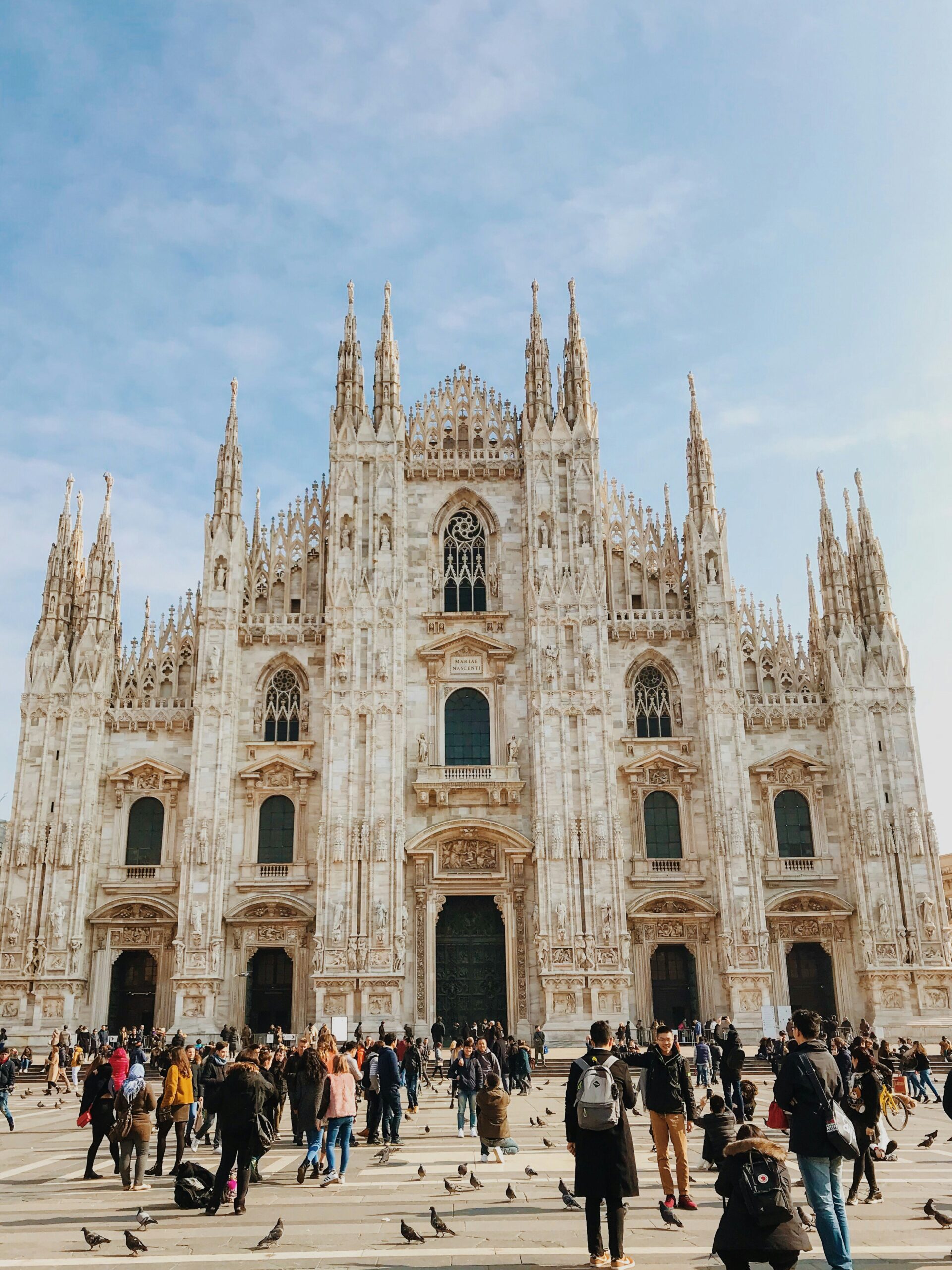 A crowd gathers in front of Milan Cathedral, showcasing its intricate Gothic architecture with numerous spires and detailed stonework, set against a clear blue sky.
