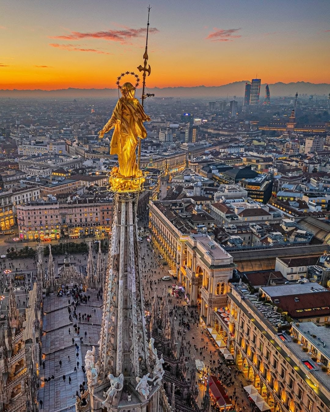 Aerial view of Milan Cathedral at sunset, featuring the golden statue of the Madonnina atop the spire, with the city skyline and crowds below.